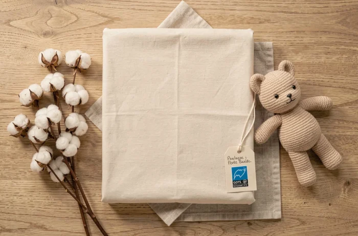 Flatlay d'un drap housse coton bio écru plié avec étiquette GOTS et accessoires naturels pour bébé sur bois clair